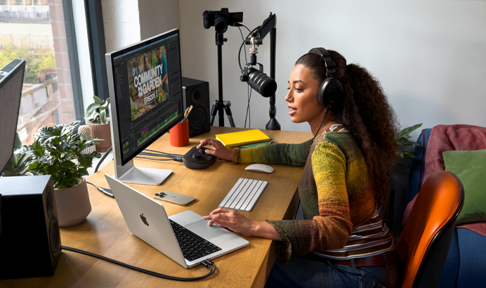 Una donna che lavora da casa seduta alla scrivania con un MacBook Pro collegato a due monitor esterni, che indossa un paio di cuffie e parla in un microfono