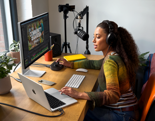 Una donna che lavora da casa seduta alla scrivania con un MacBook Pro collegato a due monitor esterni, che indossa un paio di cuffie e parla in un microfono